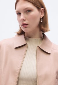 Šviesiai rožinė odinė striukė su apykakle ir priekinio užtrauktuko uždarymu, dėvima ant smulkiai briaunuoto smėlio spalvos viršaus. Lygi tekstūra, minimalistinis dizainas, laisvalaikio stilius.