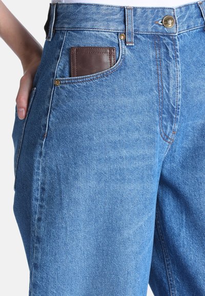 Jean bleu en denim avec un portefeuille marron partiellement visible dans la poche avant et une main reposant sur la hanche.