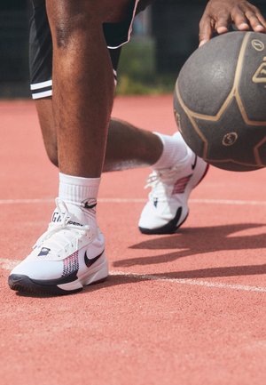 Osoba nosící bílé tenisky Nike dribluje černo-hnědý basketbal na venkovním červeném hřišti.
