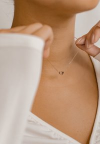 Collana con pendente a forma di cuore in argento, appesa a una delicata catena, esibita sulla pelle. Il pendente riflette la luce, mettendo in mostra una superficie liscia e lucida.