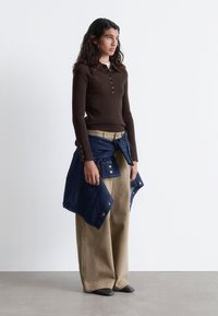 Brown ribbed long-sleeve top with button details paired with beige wide-leg pants. A blue denim jacket is tied around the waist. Black shoes.