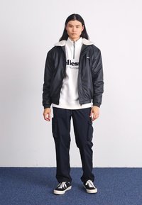Chaqueta negra de nailon con cuello de forro polar sobre un sudadero blanco con letras negras, combinada con pantalones cargo oscuros y zapatillas negras.