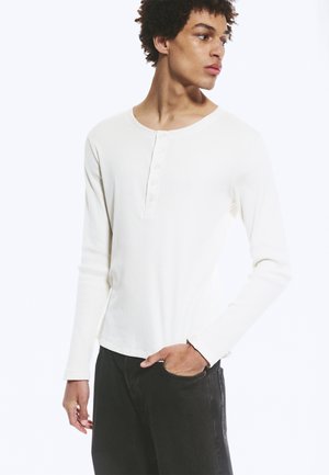 Jeune homme portant un t-shirt henley blanc à manches longues et un pantalon noir, regardant vers sa droite, une main dans la poche.