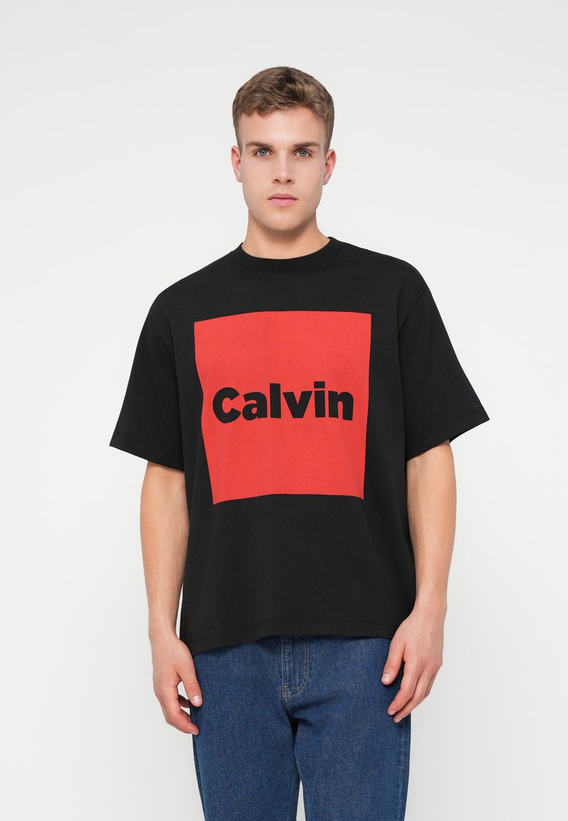 Crna pamučna majica s izrazitim crvenim kvadratom na kojem je u crnom tekstu napisano "Calvin". Kratki rukavi i opušteni kroj. U kombinaciji s plavim trapericama.