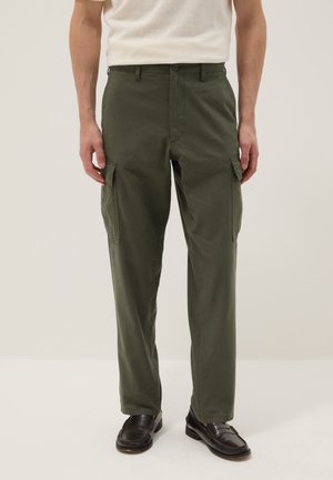 Mann trägt olivgrüne Cargohosen mit Seitentaschen, ein weißes kurzärmeliges Hemd und dunkelbraune Loafer, steht vor einem einfarbigen Hintergrund.