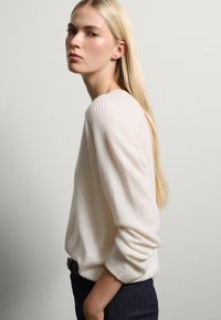 Maglione bianco a costine con maniche lunghe, realizzato in materiale morbido. Il design presenta una vestibilità comoda ed è abbinato a pantaloni scuri.