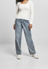 Jasnobeżowa prążkowana bluzka z długim rękawem zestawiona z luźnymi jasnoniebieskimi dżinsami i białymi tenisówkami, na tle jednolitego szarego tła.