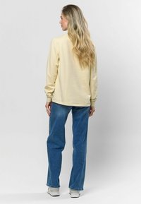 Gaiši dzeltena džemperis ar garām piedurknēm un brīvu piegriezumu, apvienots ar zilām džinsām un baltām kedām, redzams no aizmugures.