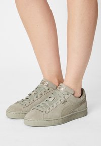Szare zamszowe sneakersy z płaskimi sznurowadłami, teksturowaną gumową podeszwą oraz srebrnym logo na języku i boku. Niskoprofilowy design z okrągłym noskiem.