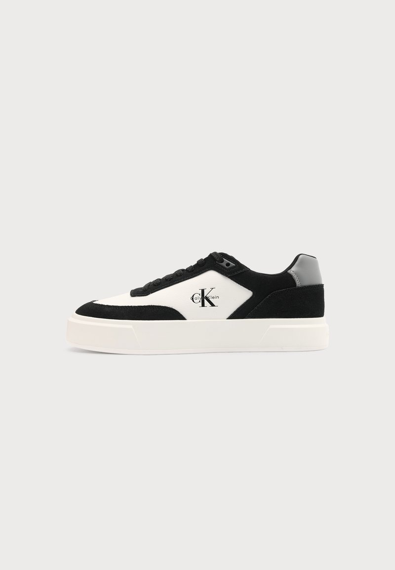 Baskets basses noires et blanches avec une épaisse semelle blanche, des lacets noirs, un œillet gris au talon, et le logo "Calvin Klein" sur le côté.