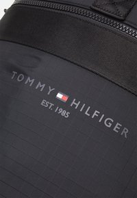 Sac à dos en tissu noir avec une surface texturée, sur lequel figure le logo "TOMMY HILFIGER" en gris et un emblème rouge-blanc-bleu. Établi en 1985.