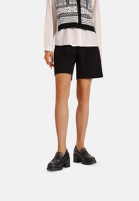 Scarpe con tacco quadrato nere con una parte superiore a motivo. Abbinato a pantaloni corto neri fino al ginocchio e a una camicia bianca con un design geometrico nero.
