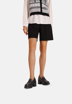 Scarpe con tacco quadrato nere con una parte superiore a motivo. Abbinato a pantaloni corto neri fino al ginocchio e a una camicia bianca con un design geometrico nero.