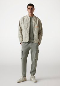 Bež vooderdatud jakk, millel on oranž logo, koos heledate roheliste spordipükstega, millel on küljetaskud ja sobiv crew neck t-särk.