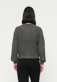 Persona con capelli scuri di lunghezza spalle, vestita con un maglione grigio lavorato a maglia e pantaloni neri, di fronte a uno sfondo bianco semplice.