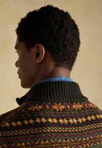 Kötött pulóver sötétzöld bordás gallérral, narancssárga, sárga és lila színű, mintákkal a hátsó részén. Világoskék ing van alatta.