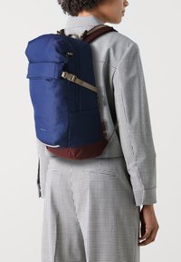 Marineblå ryggsekk med rulltopdesign, brun bunn, frontlomme og justerbare stropper foran et rutete skjorte bakgrunn.