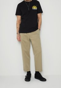 Černé tričko s krátkým rukávem a žlutým potiskem, spárováno s béžovými kalhotami rovného střihu a černými botami. Jednoduchý, casual outfit.