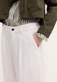 Eine Nahaufnahme einer Person in einer olivgrünen Jacke und gestreiftem Shirt, mit weißen Hosen und einer Hand in der Tasche, die ein lässiges Outfit präsentiert.