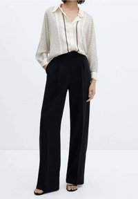 Leichte Bluse mit cremefarbenen Streifen und lockerer Passform, kombiniert mit hoch taillierten, weit geschnittenen schwarzen Hosen. Sandalen runden das Outfit ab.