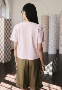 Moteris su ilgais juodais plaukais, dėvinti akinius, šviesiai rožinę marškinėlį ir alyvuogių spalvos šortus, stovinti patalpoje šalia raštuotų kolonnų.