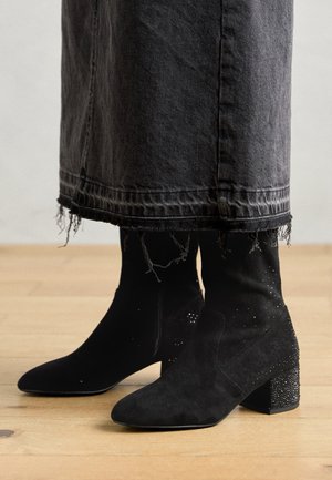 Bottines en daim noir à talons blocs et petits clous décoratifs portées avec une jupe en jean foncé à ourlet effiloché.