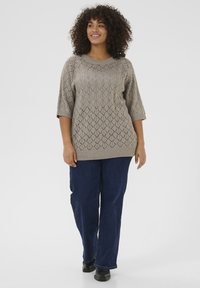 Lichtbruine, gebreide trui met een ruitpatroon, halve mouwen en een relaxte pasvorm, gecombineerd met donkerblauwe jeans met rechte pijpen.