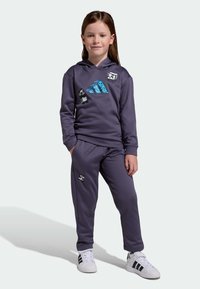 Child with long brown hair wears purple hoodie and pants with pixelated animal designs, standing with hands in pockets and white sneakers.