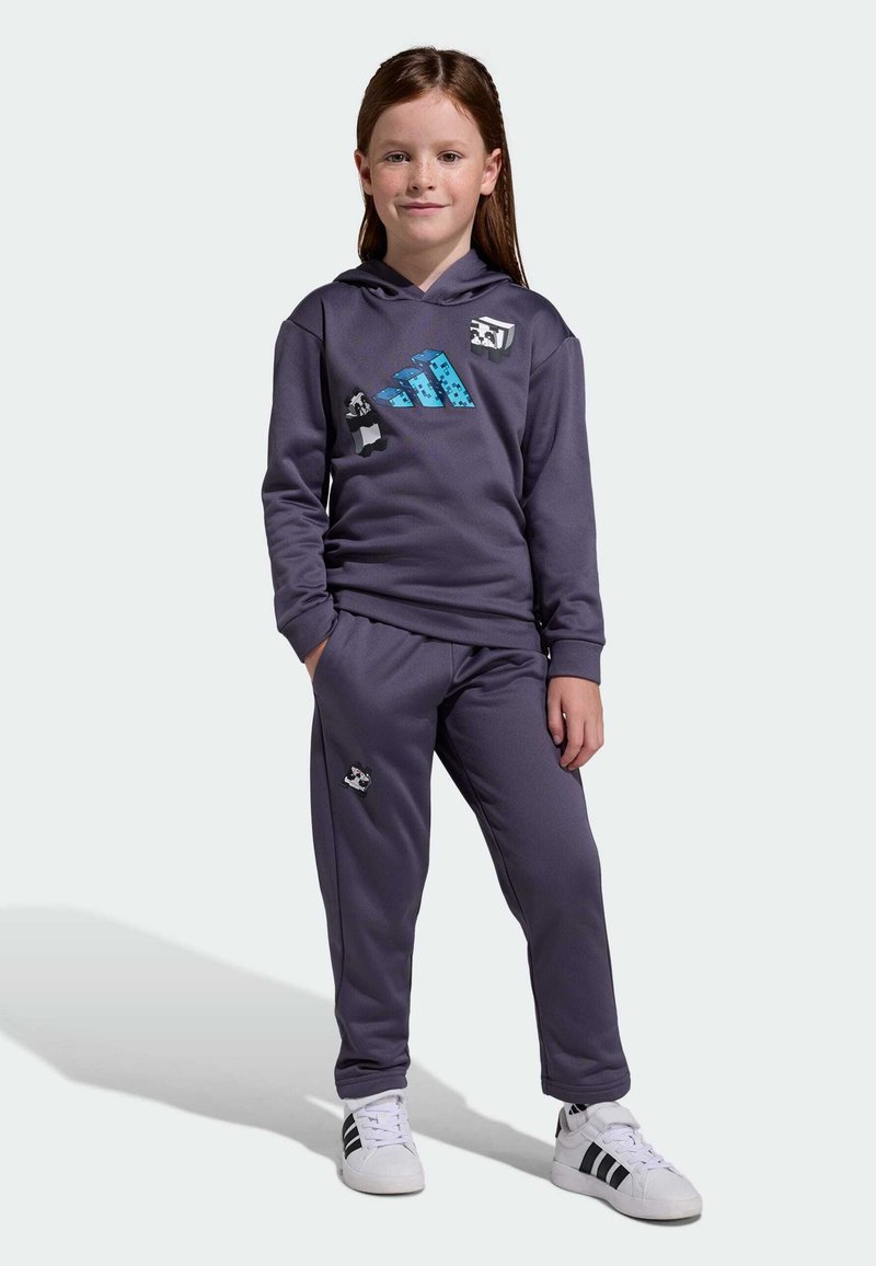 Child with long brown hair wears purple hoodie and pants with pixelated animal designs, standing with hands in pockets and white sneakers.