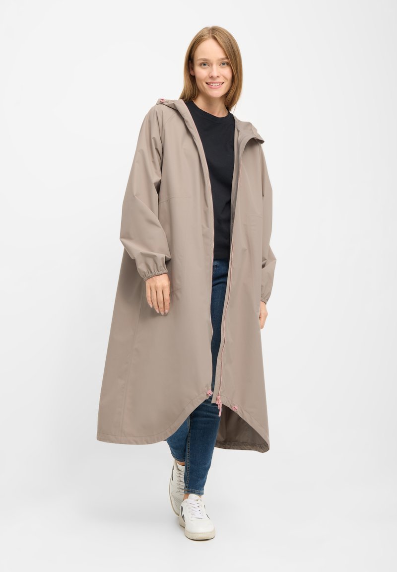 Giovane donna con impermeabile lungo beige con cappuccio, top nero, jeans blu e scarpe sportive bianche, in piedi e sorridente su sfondo bianco.