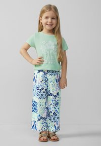 Hellgrünes T-Shirt mit floralem Grafikdesign, kombiniert mit weiten, gemusterten Hosen in Blau, Grün und Pink. Goldene Sandalen runden das Outfit ab.
