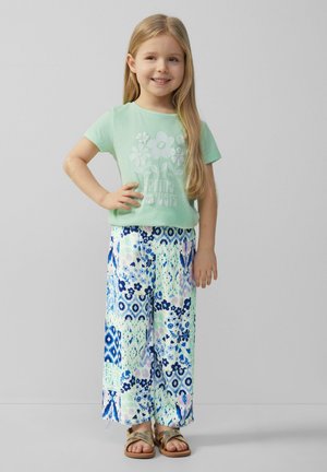 Hellgrünes T-Shirt mit floralem Grafikdesign, kombiniert mit weiten, gemusterten Hosen in Blau, Grün und Pink. Goldene Sandalen runden das Outfit ab.
