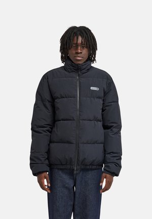 Jeune personne avec des dreadlocks foncés portant une doudoune noire et un jean bleu, debout devant un fond clair uni.