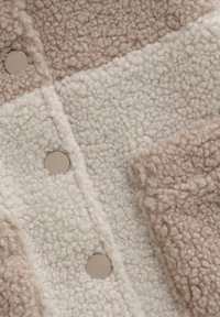 Teksturowana dzianina polarowa w beżowych i kremowych łatach; ma okrągłe beżowe guziki oraz miękką, puszystą powierzchnię o przytulnym wyglądzie.