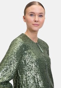 Grüne Paillettenjacke mit einem Reißverschluss, die eine strukturierte Oberfläche und eine lockere Passform aufweist. Akzente sind ein bequemer Kragen und glänzende Verzierungen.