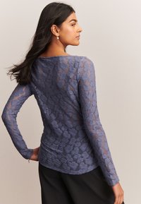 Top in pizzo a maniche lunghe in blu tenue, con motivo floreale, scollo a barca e design aderente. Tessuto morbido con dettagli trasparenti.