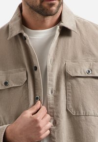 Beige button-up shirt with two chest pockets, made of textured fabric. Features grey buttons and a classic collar. Worn over a white shirt.