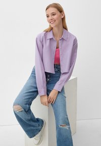 Korte lavendelkleurige cropped button-up shirt, gecombineerd met vervaagde blauwe flared jeans met gescheurde details en witte sneakers. Eenvoudige achtergrond.