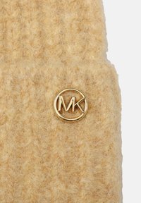 Knit wool beanie in soft beige, featuring a textured pattern. Gold circular logo accenting the front enhances the design.