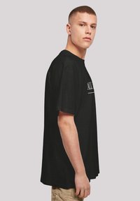 Schwarzes Baumwoll-T-Shirt mit lockerer Passform, kurzen Ärmeln, rundem Halsausschnitt und weißem Textdetail auf der Vorderseite. Einfarbiger Rücken.