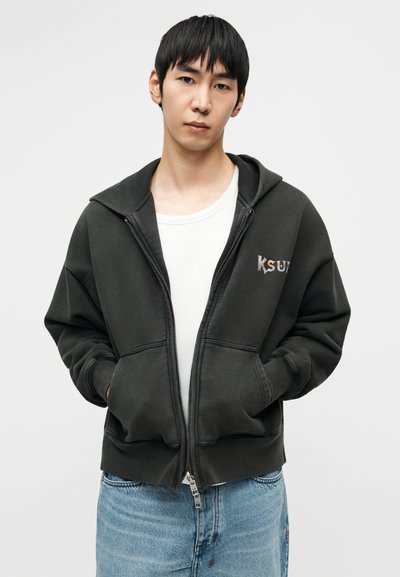 Σκούρο πράσινο φούτερ με φερμουάρ και μεγάλο κουκούλι, μπροστινές τσέπες και κεντημένο λογότυπο "KSUT". Φορεμένο πάνω από ένα λευκό μπλουζάκι και ανοιχτόχρωμα μπλε τζιν.
