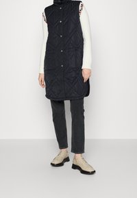 Gesteppte, ärmellose schwarze Weste mit hohem Kragen, vorderen Taschen und Knopfverschluss. Kombiniert mit dunklen Jeans und beigen Schuhen.