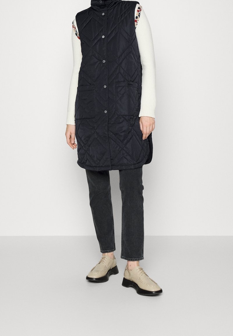 Gesteppte, ärmellose schwarze Weste mit hohem Kragen, vorderen Taschen und Knopfverschluss. Kombiniert mit dunklen Jeans und beigen Schuhen.