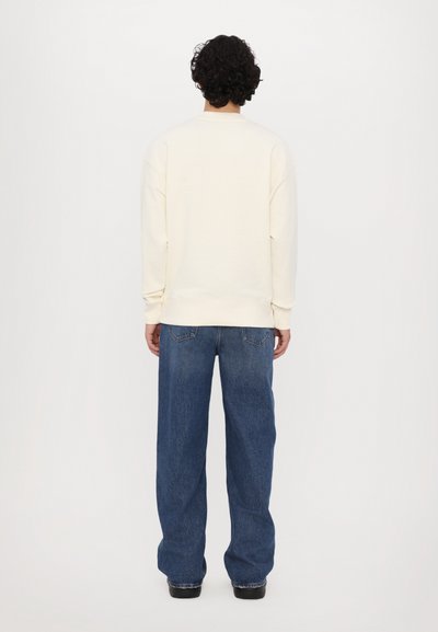 Sweatshirt à manches longues couleur crème avec un col rond, associé à un jean en denim bleu ample. Le tissu semble doux et offre une coupe décontractée.
