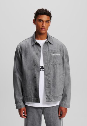 Giacca in denim grigia con vestibilità rilassata, chiusura a bottoni e taschino sul petto. Indossata sopra una t-shirt bianca con testo nero.