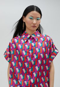 Short-sleeve shirt in smooth satin with a geometric pattern of pink, red, and teal shapes. Features a pointed collar and button-down front.