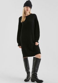 Vrouw in een zwarte gebreide jurk, zwarte beanie, en hooglopende zwarte leren laarzen staande tegen een effen lichte achtergrond.