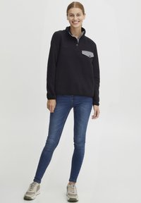 Sort fleece pullover med en grå brystlomme, krave-design, lange ærmer, og båret med blå skinny jeans og lyse sneakers.