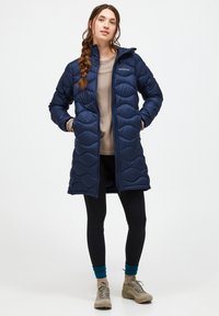 Marineblauwe pufferjas met een gequilt patroon, ritssluiting en capuchon. Gedragen over een beige top en zwarte leggings, gecombineerd met tan kleurige wandelschoenen.