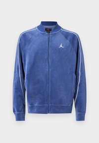 Casaco azul com fecho éclair, feito de material suave. Apresenta acabamentos brancos nas mangas e um logótipo no peito. Gola e punhos canelados.